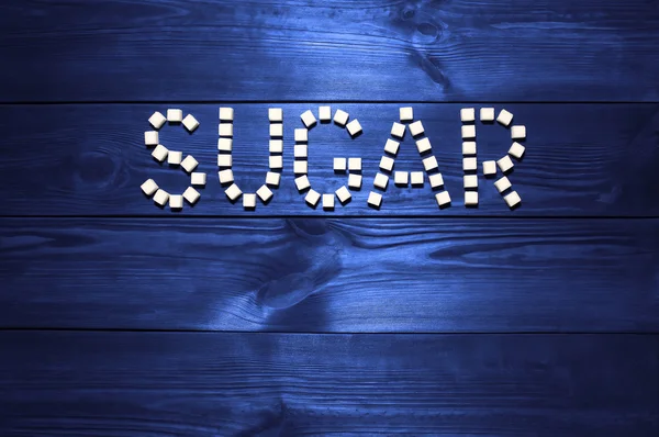 The word \'sugar\', built from white cubes pieces of sugar on background  of panel from vintage wooden boards dark blue color in high contrast dramatic lighting  \'Hard light\'