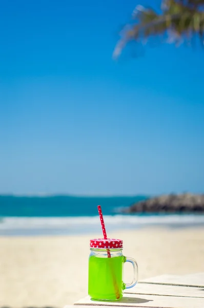 Vertical photo of green juice in a funny glass with a red straw, beach as background