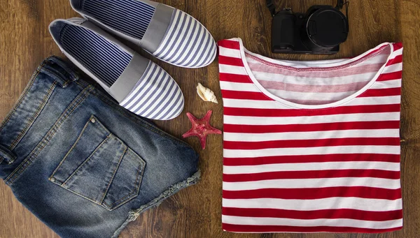 Set clothes for going to sea: jeans shorts, a striped shirt and striped sneakers, photocamera, shells, a top view of a wooden background