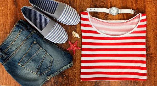 Set clothes for going to sea: jeans shorts, a striped shirt and  sneakers, watches, shells,  top view of  wooden background