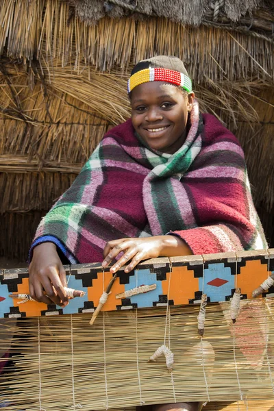 Smiling beautiful African Zulu woman in ethnic clothes weaves straw carpet.