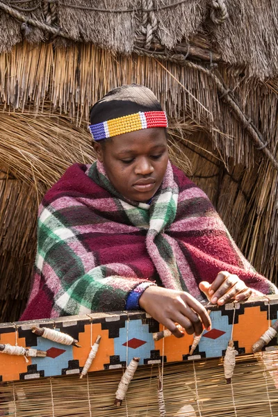 Beautiful African Zulu woman in ethnic clothes weaves straw carpet.