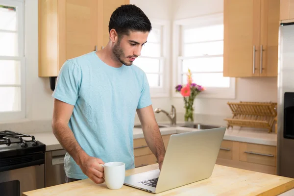 Handsome young man with laptop computer and coffee at home in kitchen typing working networking internet
