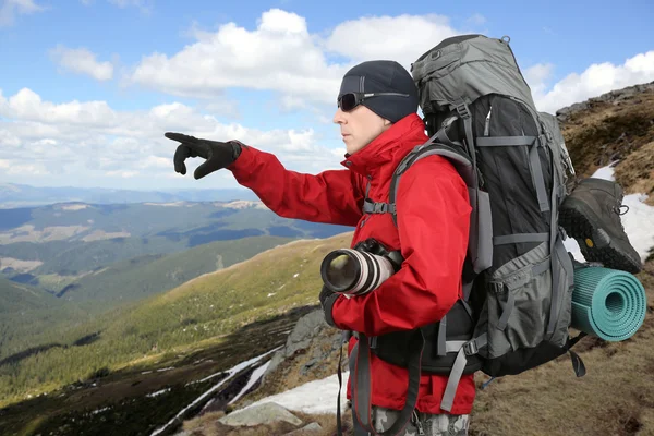 Traveler in red jacket with backpack and camera in hand points into the distance hand being on the hillside