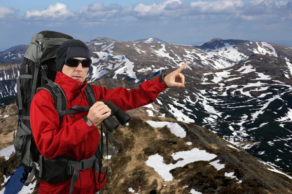 Traveler with backpack red jacket with binoculars in hand on the slopes of mount points into the distance with your finger
