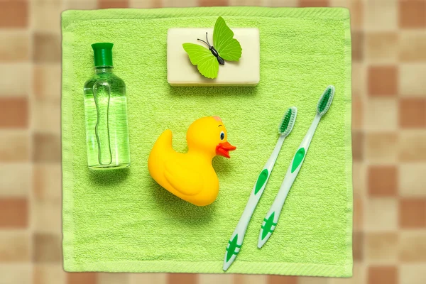 Bathroom accessories on a green towel. Rubber ducky, toothbrushes, soap and lotion. Baby care accessories for bath. Yellow duckling toy for kids.