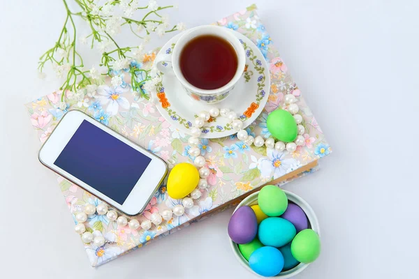 Phone with a cup of tea and pearl necklace with flowers on a white satin background. A book, tea cup, telephone and decoration. Tea time and easter painted eggs