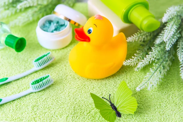 Bathroom accessories on a green towel. Rubber ducky, toothbrushes, sea salt, soap and lotion. Baby care accessories for bath. Yellow duckling toy for kids.
