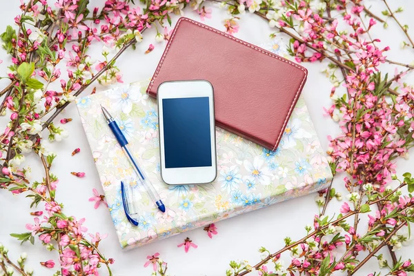 Office accessories with a beautiful blossom flower branches on a white background. Phone, notebook and a pen with a book.