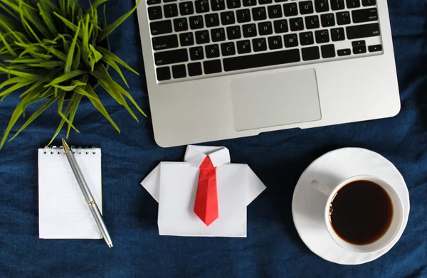 Laptop keyboard, white cup of tea on saucer,white origami shirt with red tie, notepad, pen and green plant in the corner on dark blue crumpled jeans background.