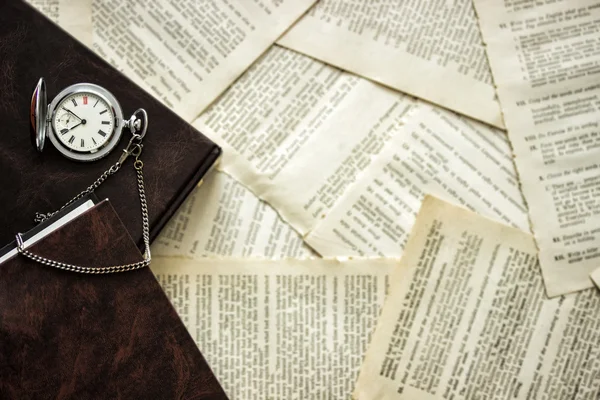 Old silver pocket watch and a book lying on the table covered with book pages. Top view