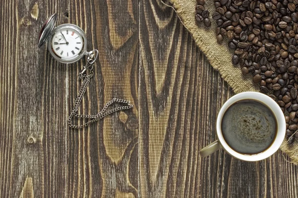 A pocket watch with a cup of coffee and crema is in the corner of a wooden table on a burlap canvas close to crumble coffee beans. Close up. Top view