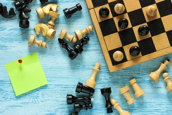 Old cropped chessboard with black and white figures and green sticker on blue wooden background. Top view. Flat lay.