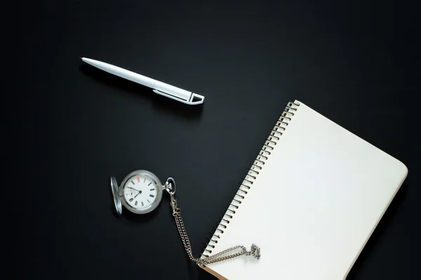 Single simple empty white notebook with a blank for drawing or writing and white plastic pen and silver pocket watch are on a Desktop from black chalkboard. Top view. Mockup. Flat lay