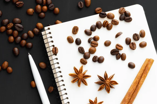 Spilled coffee beans, cinnamon sticks, cardamom few flowers, white plastic pen and a blank notebook on a black background. Top view. Mock up. Flat lay