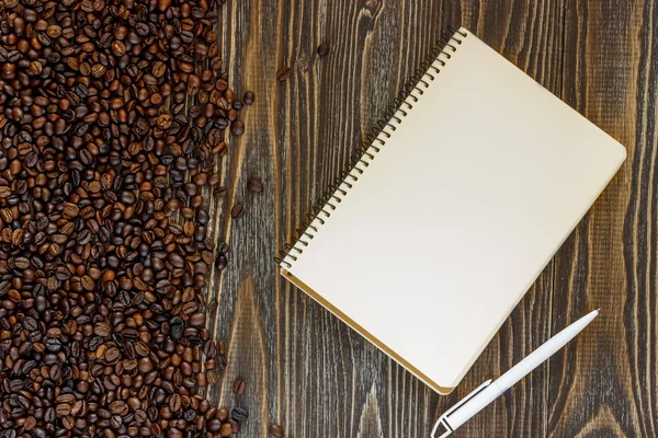 Single simple empty white notebook with a blank for drawing or writing, white plastic pen and scattered roasted coffee beans are on a Desktop from wooden plank. Top view. Mock up. Flat lay