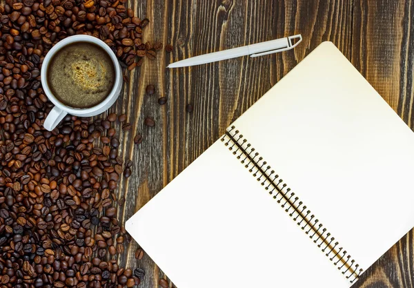 Cup of coffee single simple empty white notebook with a blank for drawing or writing, white plastic pen and scattered roasted coffee beans are on a Desktop from wooden plank. Top view. Mock up. Flat l