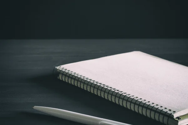 Single simple empty white notebook with a blank for drawing or writing and white plastic pen are on a Desktop from black chalkboard. Mockup. Toned and grained
