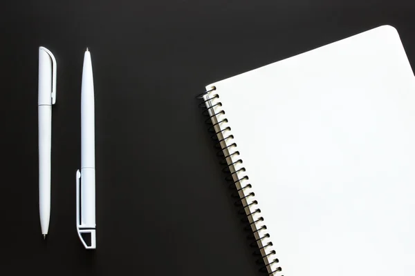 Single simple empty white notebook with a blank for drawing or writing and white plastic pen are on a Desktop from black chalkboard. Top view. Mockup. Flat lay
