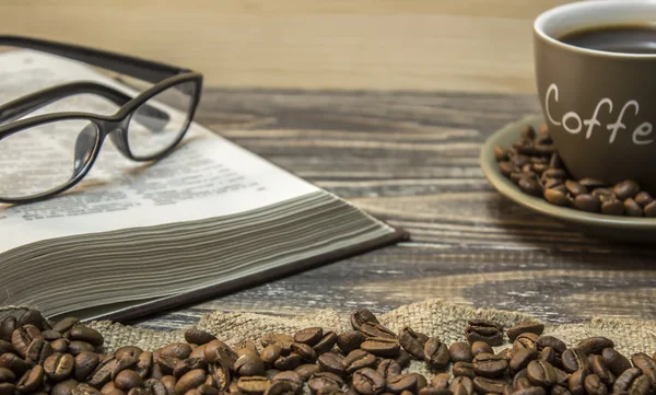 A cup of coffee with scattered coffee beans and open book with eyeglasses are  stands on a piece of burlap. Focus on beans on burlap