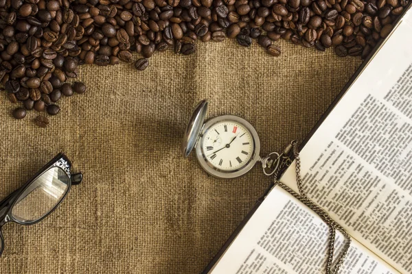 Background of roasted coffee beans scattered on the canvas of burlap, glasses, open book and pocket watches