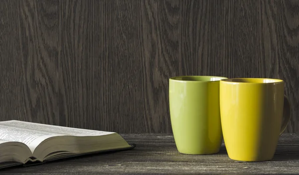 Two ceramic mugs yellow and green stand in the corner of a wooden table in front of an open book