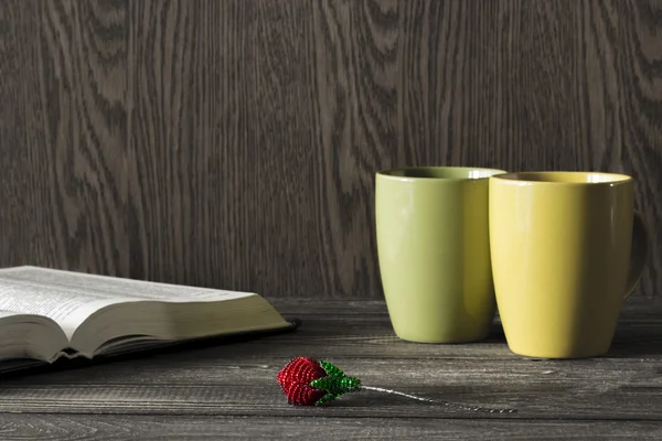 Bright artificial rose bead lies in front of the yellow and green mug with open book on a wooden table. Focus on flower