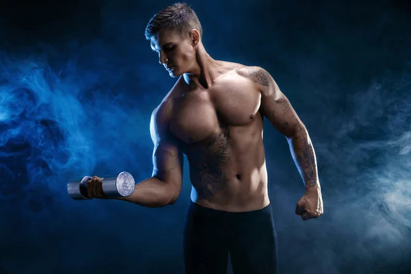 Closeup of a handsome power athletic man bodybuilder doing exercises with dumbbell. Fitness muscular body on dark  smoke background. Perfect male. Awesome bodybuilder, tattoo, posing.