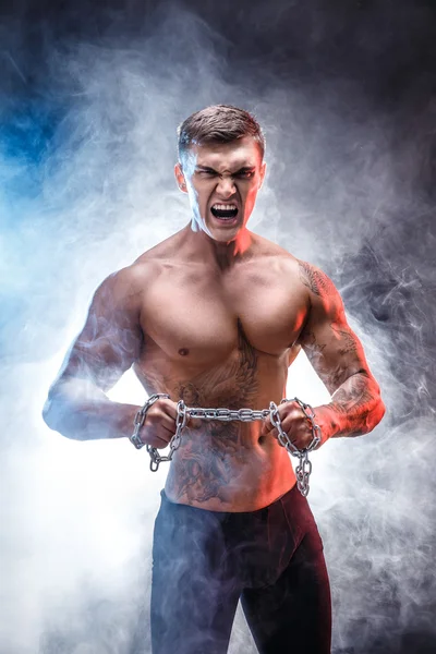 Handsome power athletic man bodybuilder doing exercises with chain, tearing. Fitness muscular body on dark background. Perfect male. Awesome bodybuilder, tattoo, posing.