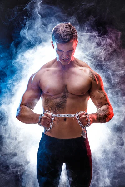 Handsome power athletic man bodybuilder doing exercises with chain, tearing. Fitness muscular body on dark background. Perfect male. Awesome bodybuilder, tattoo, posing.