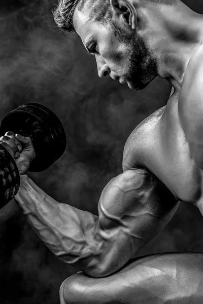 Closeup of a handsome power athletic man bodybuilder doing exercises with dumbbell. Fitness muscular body on dark smoky background. Selective Focus. Awesome bodybuilder, posing. Black and white.