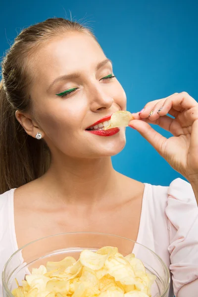 Portrait of fashionable young blond woman in pink-red dress, beautiful lips, bright make-up holding, eating fried potato, fries, chips and posing over blue background. Unhealthy eating. Junk food