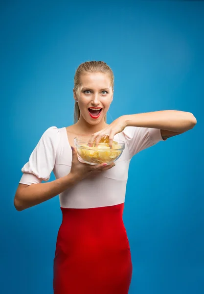 Portrait of fashionable young blond woman in pink-red dress, beautiful lips, bright make-up holding, eating fried potato, fries, chips and posing over blue background. Unhealthy eating. Junk food