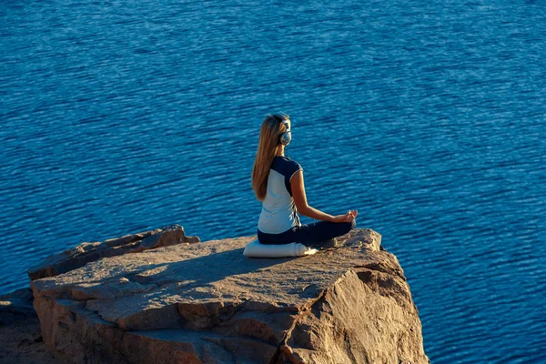 Woman wearing sport suit and headphones Sitting in Lotus Position on Rock Above the Sea, Meditating, listen music. Yoga Outdoor.