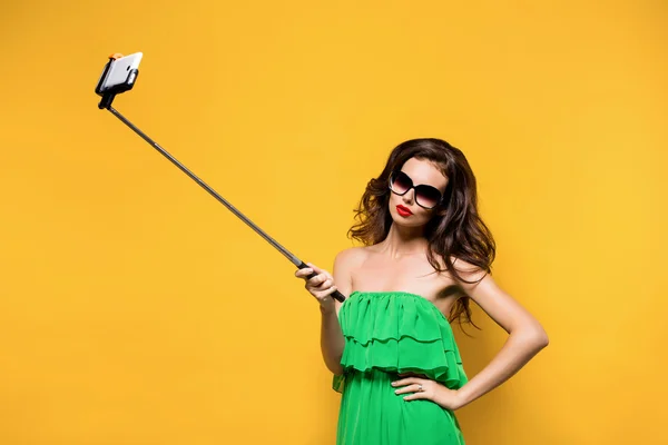 Portrait of young model in green dress and sunglasses posing while making selfie against yellow background.