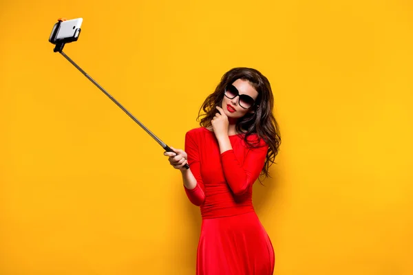 Portrait of beautiful brunette in awesome dress and red lips wearing sunglasses while making selfie with stick. Isolated