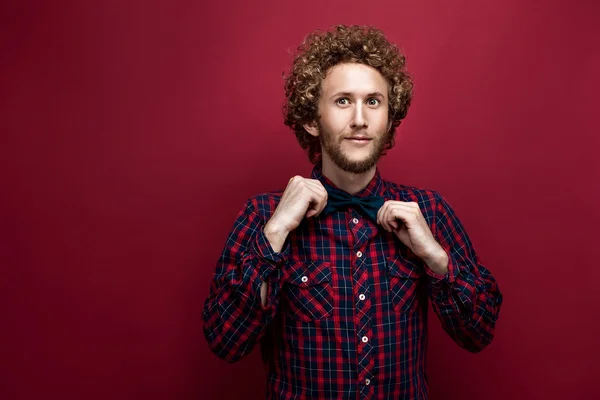 Portrait of surprised curly-haired man in checked shirt and bow-tie on red background. Isolate