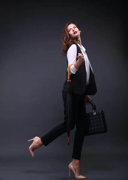 Pretty female business lady in a black jacket white shirt, umbrella, handbag on a dark background. Studio shot.