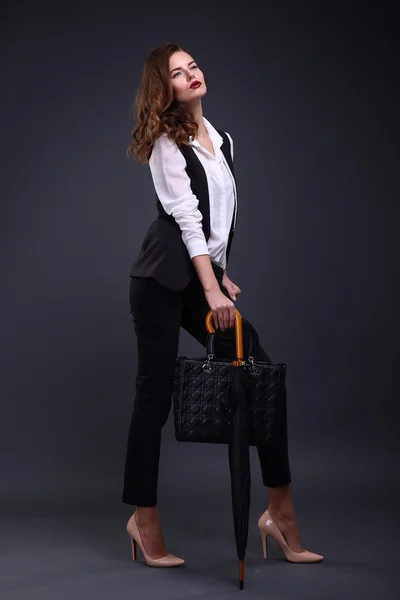 Pretty female business lady in a black jacket white shirt, umbrella, handbag on a dark background. Studio shot.