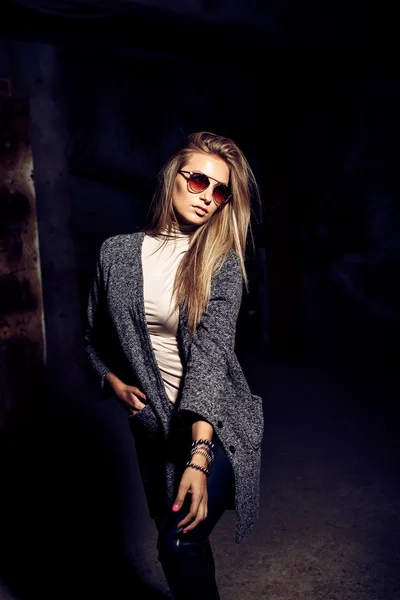 Portrait of a beautiful young blond womanin grey coat, sunglasses, leather pants in an industrial background. Abandoned metal factory.