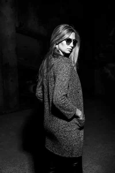 Portrait of a beautiful young blond womanin grey coat, sunglasses, leather pants in an industrial background. Abandoned metal factory. Black and white photo.
