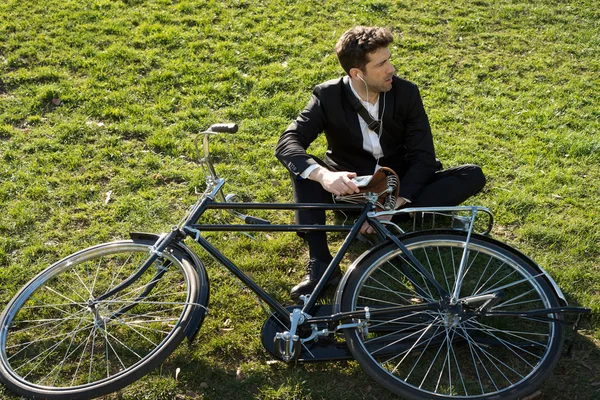 Businessman with bicycle listens to earpieces seated in the park