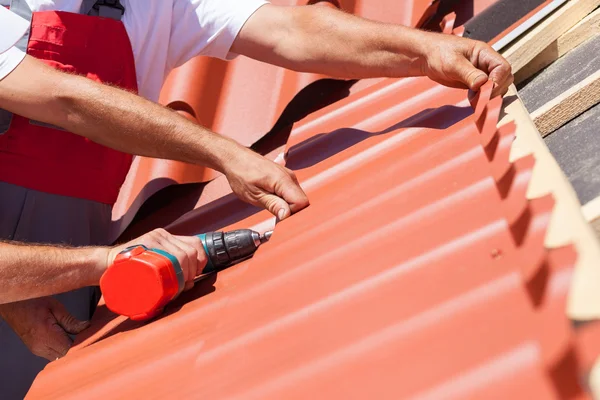 Workers on a roof with electric drill installing red metal tile on wooden house