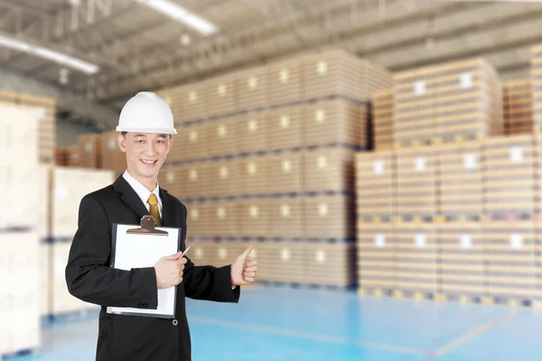 Happy asian young man in suit with helmet and clipboard gesturing thumbs up in warehouse