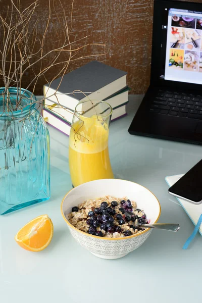 Breakfast with blueberry muesli, orange juice on glass table, books, laptop on background.
