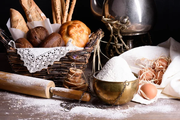 Variety of bread and sweet pastry, flour, eggs and rolling pin. Wooden table, dark background.