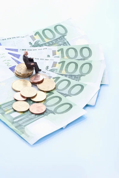 Male figurine sitting on stack of euro coins with 100 euro notes