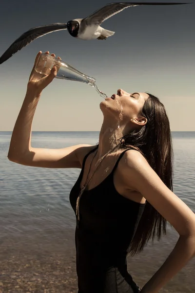 Young and sexy brunette holding bottle, drinking water in a black wet swimsuit on the sea beach, gull is flying nearby and watching her