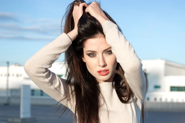 Attractive young brunette woman with pretty makeup shaking head, touching hair by her hands, standing outdoors on a bright sunny day in front of buildings under the blue sky