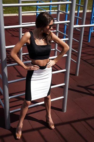 Young sporty teenage model making crossfit exercises and posing on playground near horizontal and parallel bars, stairs, wearing black and white top and skirt